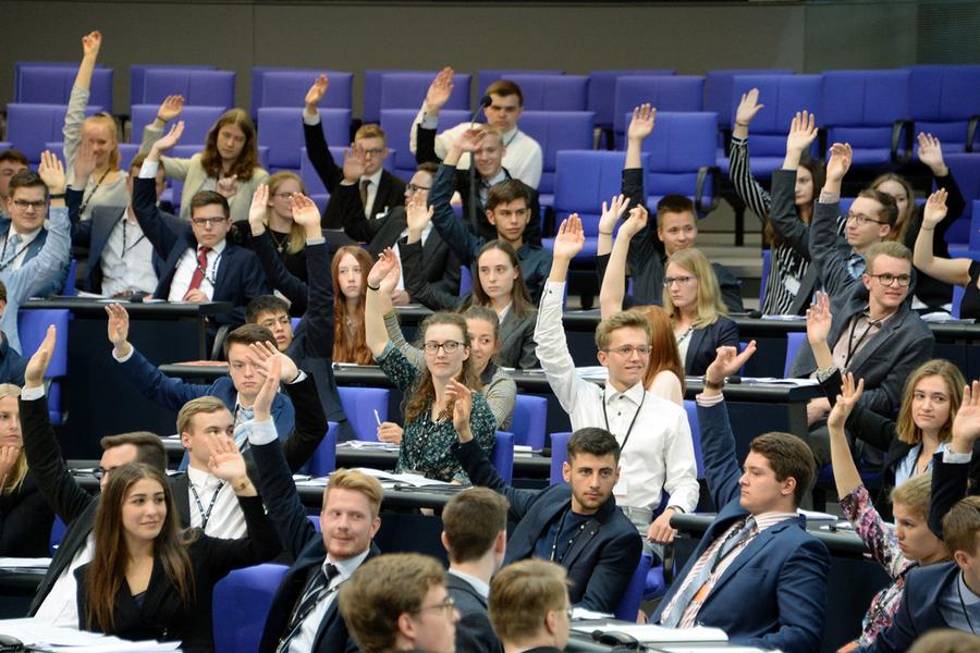 Viele Personen sitzen in einem Plenum und viele davon heben zur Abstimmung einen Arm.