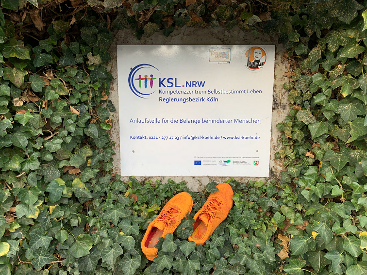 orangenes Schuhpaar vor KSL Köln Schild